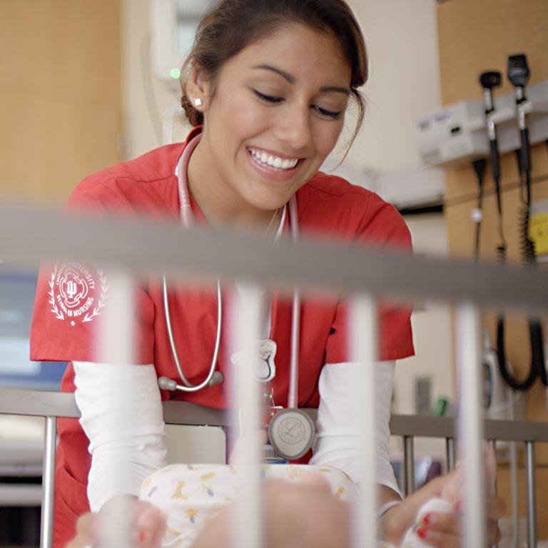 A nurse in red scrubs leans over the edge of a crib to play with a baby. 